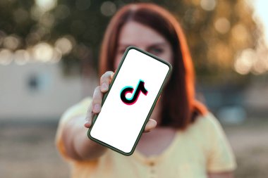 Odessa, Ukrayna, 6 Eylül 2020: Genç kız parktaki yeşil çimenlerde oturan Tiktok logosu yazılı cihaz kullanıyor