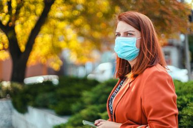 Young girl in a medical mask and suit holds a smartphone in her hands and looks to the side on the street. Coronavirus, covid 19 concept clipart