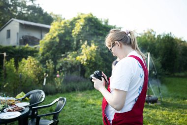 Kırmızı önlüklü hamile bir kadın bahçede dikiliyor ve elinde fotoğraf makinesi, ekrandaki resimlere bakıyor. Annelik yaşam tarzı, hamilelik süresince hobiler ve aile eğlencesi kavramı. Beklentinin sembolü