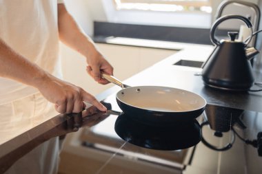 Modern mutfakta, beyaz elbiseli bir adamın elektrikli ocağa tava koyduğu yakın plan. Yemek pişirme, günlük rutin ve ev hayatı kavramı. Hazırlık, rahatlık ve minimalist iç mekan sembolü