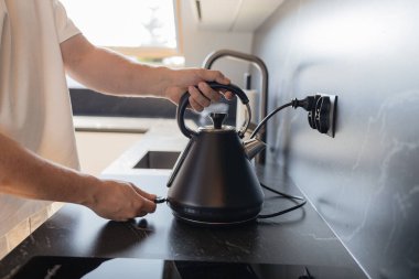 Modern mutfaktaki mermer duvarlı ocağa siyah elektrikli su ısıtıcısı koyan adamın yakın çekimi. Ev hayatı, yemek pişirme ve günlük rutin şeyler. Ev konforunun sembolü, şık iç mekan ve