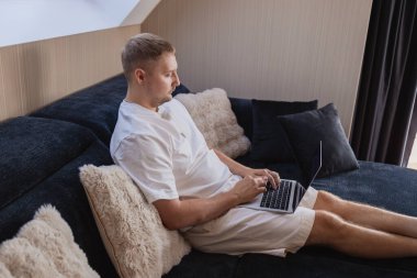 Dizüstü bilgisayarı ve akıllı telefonuyla kanepede oturan genç adam uzaktan çalışıyor ya da online işleri yönetiyor. Serbest yaşam, dijital yaşam tarzı, mobil teknoloji, e-bankacılık, online alışveriş, iletişim kavramı