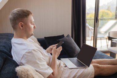 Dizüstü bilgisayarı ve akıllı telefonuyla kanepede oturan genç adam uzaktan çalışıyor ya da online işleri yönetiyor. Serbest yaşam, dijital yaşam tarzı, mobil teknoloji, e-bankacılık, online alışveriş, iletişim kavramı