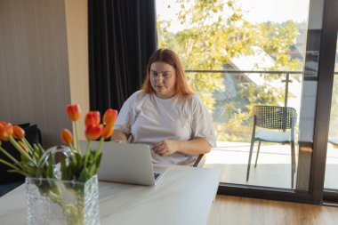 Masasında dizüstü bilgisayarıyla oturan genç bir kadın ev içinde uzaktan çalışıyor ya da internetten ders çalışıyor. Serbest yaşam, dijital yaşam tarzı, uzak çalışma, e-öğrenme, iletişim, modern teknoloji kavramı