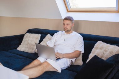 Dizüstü bilgisayarı ve akıllı telefonuyla kanepede oturan genç adam uzaktan çalışıyor ya da online işleri yönetiyor. Serbest yaşam, dijital yaşam tarzı, mobil teknoloji, e-bankacılık, online alışveriş, iletişim kavramı