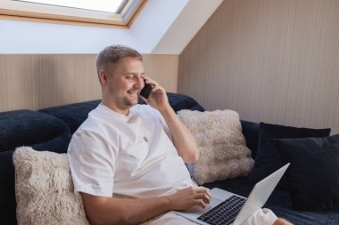 Kanepede dizüstü bilgisayarla oturan ve akıllı telefondan konuşan gülümseyen adam. Uzaktan çalışma, çevrimiçi iletişim, serbest çalışma, dijital yaşam tarzı, e-bankacılık, online alışveriş veya iş görüşmesi kavramı. Samimi