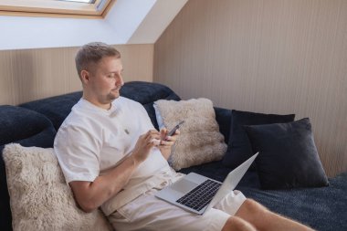 Dizüstü bilgisayarı ve akıllı telefonuyla kanepede oturan genç adam uzaktan çalışıyor ya da online işleri yönetiyor. Serbest yaşam, dijital yaşam tarzı, mobil teknoloji, e-bankacılık, online alışveriş, iletişim kavramı