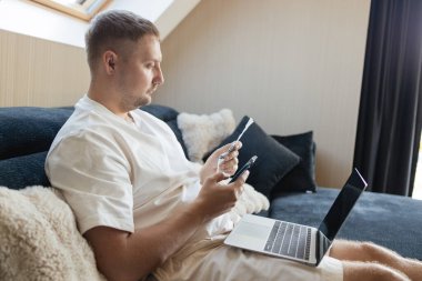 Genç adam bilgisayar ve telefonla masada oturuyor, faturaları ödüyor ya da internetten alışveriş yapıyor. Nakitsiz ekonomi, mobil bankacılık, ev bütçesi, mali teknoloji ve uzak çalışma.