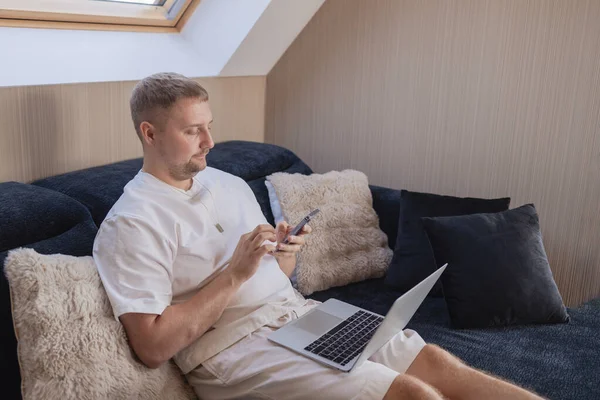 Dizüstü bilgisayarı ve akıllı telefonuyla kanepede oturan genç adam uzaktan çalışıyor ya da online işleri yönetiyor. Serbest yaşam, dijital yaşam tarzı, mobil teknoloji, e-bankacılık, online alışveriş, iletişim kavramı