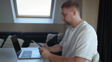 Evde oturan genç adam, güvenli online ödeme için dizüstü bilgisayar, akıllı telefon ve kredi kartı kullanıyor. Günlük yaşamda dijital bankacılık, uzaktan çalışma, finansal planlama ve modern teknoloji kavramı.
