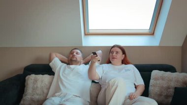 Tavan penceresinin altında rahat koltukta oturan genç bir çift birlikte rahatlıyor ve uzaktan kumandayla televizyon seyrediyor. Boş zaman kavramı, aile hayatı, rahatlık, hafta sonu rahatlığı, ev eğlencesi.