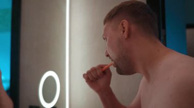 Bir adam banyo aynasının önünde dişlerini fırçalıyor, modern ışıkla aydınlatılıyor. Videoda sabah temizliği alışkanlıkları, kişisel bakım ve sağlıklı yaşam tarzları vurgulanıyor.