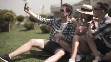 Modern bir şehirde çimlerin üzerinde oturmuş, farklı ülkelerin başkentlerini gezerken üç arkadaş selfie çekiyor. Seyahat konsepti. Arkadaş kavramı. Prores 422