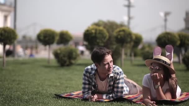 Dos yong en el amor estudiantes pasar tiempo acostado en la hierba verde cerca de la orilla del río pasando el rato en una ciudad esperando a un amigo para unirse a ellos. Pareja guapa pasar tiempo juntos en el fondo urbano de la ciudad — Vídeos de Stock