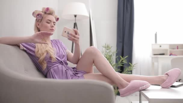 Блондинка робить селфі з завитими волоссям, сидячи вдома на дивані в фіолетовому халаті. Залишайтеся вдома. Концепція краси. Пропозиції 422 — стокове відео