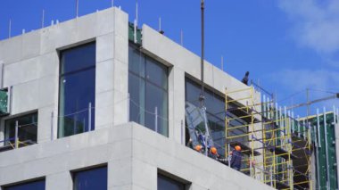İnşaatçılar yeni inşa edilmiş bir binanın camını cilaladılar. En üst kata koymak için vinç tarafından kaldırılan camı tutuyorlar. Nisan 2020. Kiev, Ukrayna. Prores 422