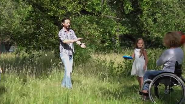 Família com uma mãe em cadeira de rodas brincando com crianças ao ar livre jogando bola e frisbee. Mãe e pai com filhos em piquenique. Conceito de pessoas com deficiência. Conceito de retrato de família. Prores 422 — Vídeo de Stock