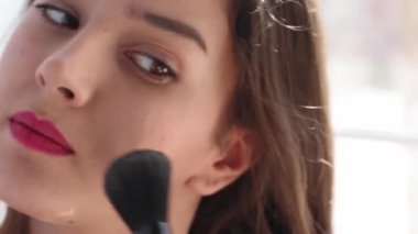 Elinde fırça ve elinde aynayla pencerenin yanında duran makyajcı bir kızı giydirmek. Evde kal konsepti. Güzellik konsepti. Prores 422