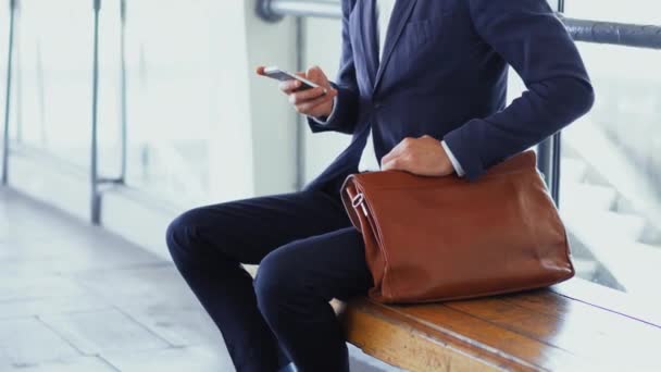 Freelancer con smartphone en la mano y traje de negocios se sienta en el banco en la estación de metro, esperando a que su tren llegue a la oficina. Hombre de negocios con un maletín en el metro. De cerca. Prores 422 — Vídeos de Stock