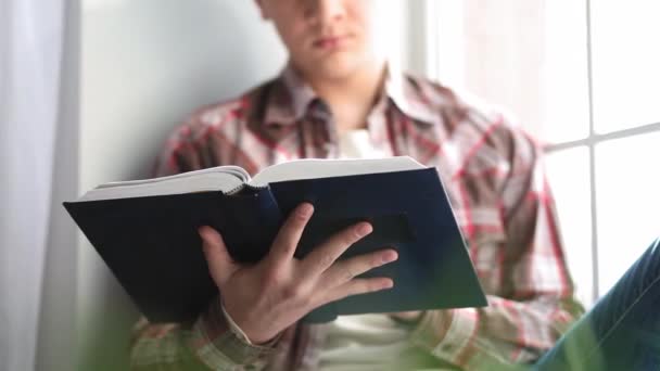 Estudante cara com um livro em mãos senta-se em um peitoril da janela se preparando para uma palestra ou exames. Aprendizagem à distância durante o período de quarentena. Conceito de educação. Feche o foco seletivo no livro. Prores 422 — Vídeo de Stock