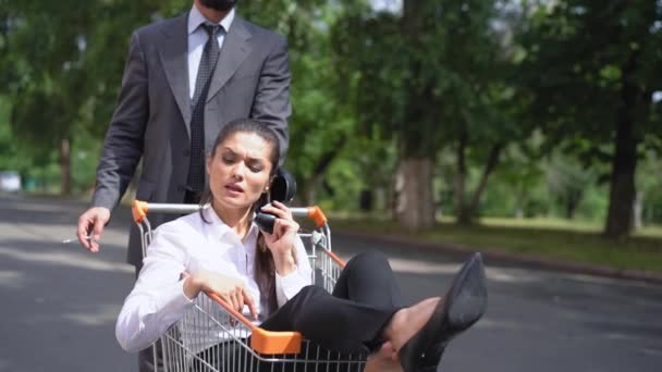 Hombre en traje de negocios empujar mujer freelancer que ha perdido la cabeza hablando en un zapato como en el teléfono en la calle en un carrito de la compra mientras fuma cigarrillo. Acoso en el trabajo. Prores 422 — Vídeo de stock