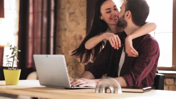 Egy fiatal szabadúszó dolgozik egy laptop mögött, miközben otthon ül. A felesége hirtelen megöleli és gyengéden megcsókolja. Családi fogalom. Gyümölcsfélék — Stock videók