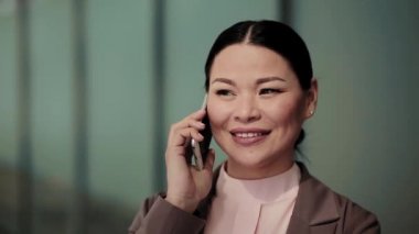 Asyalı iş kadını dışarıda ofis binasının yanında telefonla konuşuyor. Müşteriyle telefonda konuşan güzel bir kadın. İş konsepti. Kapatın. Tonlu görüntüler. Prores 422