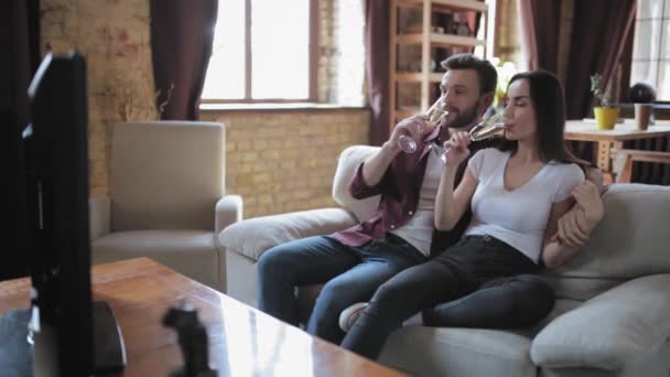 Los jóvenes celebran el aniversario tomando una copa de champán mientras están sentados en un sofá en una sala de estar frente al televisor viendo el día de su boda. Concepto familiar. Prores 422 — Vídeos de Stock