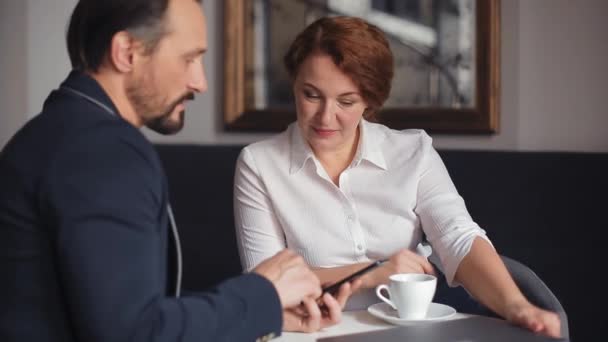 1 Üzleti férfi öltönyben bemutató kolléga nő projekt információ frissítések okostelefon, miközben beszél ül egy székben egy hangulatos kávézó csésze kávét. Gyümölcsfélék — Stock videók