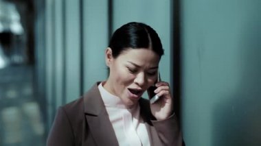 Ofis binasının yanında Asyalı iş kadını yürüyor ve telefonda konuşuyor. Müşteriyle telefonda konuşan güzel bir kadın. İş konsepti. Soğuk tonda. Tonlu görüntüler. Prores 422