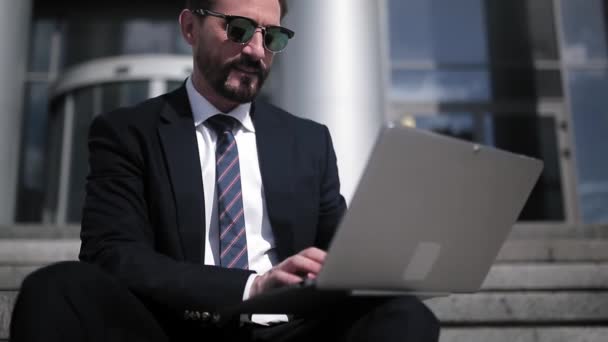Zakenman werkend op laptop zittend op een trap in de buurt van kantoorgebouw in zakenpak en zonnebril. Koffiepauze voor zakenmensen. Bedrijfsconcept. Prores 422 — Stockvideo