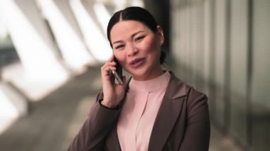 Asyalı iş kadını dışarıda ofis binasının yanında telefonla konuşuyor. Güzel bir kadın iş arkadaşıyla telefonda konuşuyor. İş konsepti. Tonlu görüntüler. Prores 422