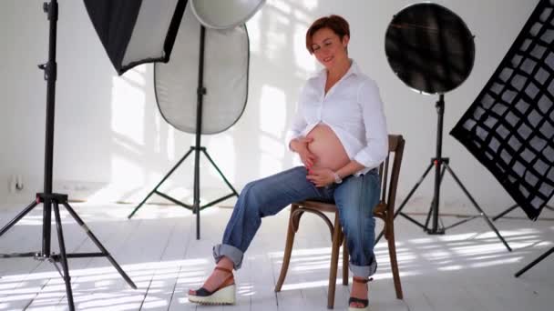 Mujer embarazada con camisa blanca masajeando su vientre sentada en un estudio con equipo reflector vista de cuerpo completo. Mujer joven con el pelo corto esperando un bebé. De cerca. Prores 422 — Vídeo de stock