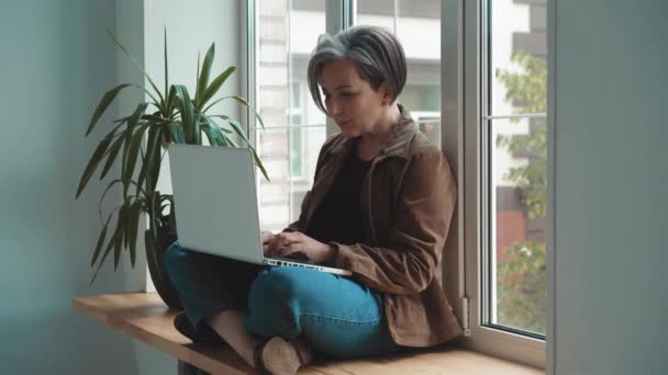 Gray haired woman working on laptop sitting leaned on the window. Freelancer woman found a free quiet place and works sitting in a Turkish pose. Business concept. Prores 422 — Stock Video