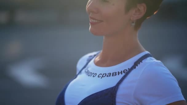 Mujer embarazada de pie junto a la carretera en overoles de mezclilla y camiseta blanca masajeando su vientre. Mujer joven con el pelo corto esperando un bebé. Cerrar la diapositiva de la cámara hacia abajo. Prores 422 — Vídeos de Stock