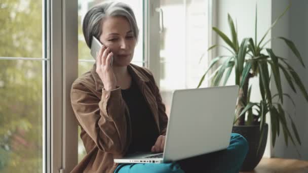 Busy freelance sales woman sitting at office window and using her laptop while talking on mobile. Businesswoman working online. Prores 422 — Stock Video