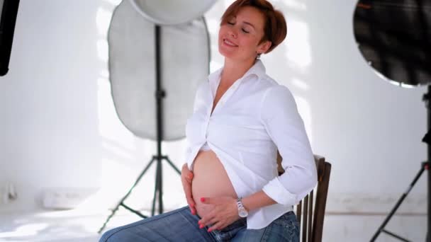 Mulher grávida feliz tendo sessão de fotos em um estúdio branco com equipamentos de luzes vestindo camisa branca massageando sua barriga. Jovem se preparando para se tornar uma mulher mãe. Fecha. Prores 422 — Vídeo de Stock