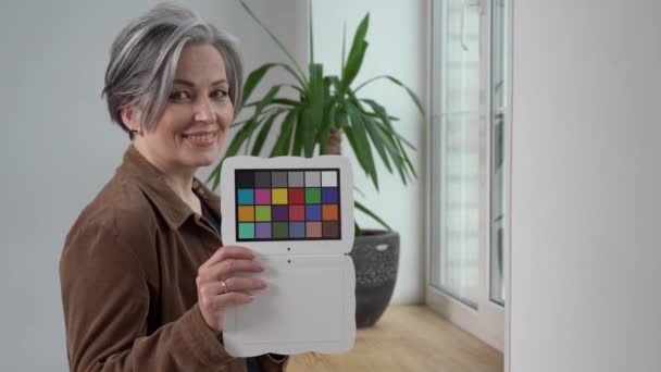 Madura mujer de negocios de pelo gris sostiene el inspector de color sonriendo a la cámara. Mujer con chaqueta marrón muestra el objetivo de ajuste de color de pie junto a la ventana. Concepto de fotografía. Prores 422 — Vídeos de Stock