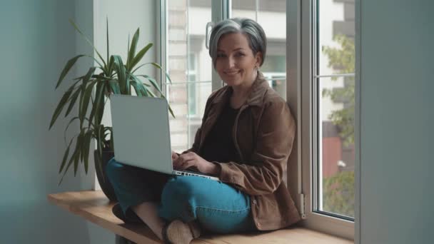 Glad mogen frilansare gråhårig kvinna som arbetar på laptop sitter lutad på fönstret. Kvinna ler mot kameran på en lugn plats och arbetar i en turkisk pose. Affärsidé. Prores 422 — Stockvideo