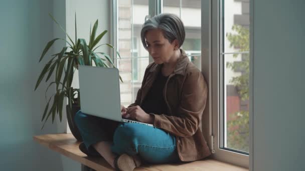 Mature businesswoman working on laptop in her workstation. Businesswoman looking at camera and smile working on laptop. Gray-haired aged woman sits cross-legged on wide windowsill. Prores 422 — Stock Video