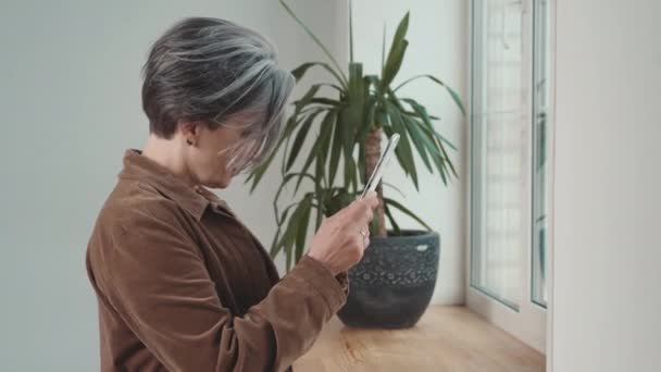 Femme mature fixant ses cheveux gris à l'aide d'une tablette numérique. Femme utilisant la caméra frontale tablette comme miroir habillé de façon décontractée debout à la maison ou au bureau à l'intérieur. Prores 422 — Video