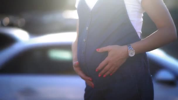 Mulher de meia-idade com cabelo curto esperando um bebê. Mulher grávida em pé ao lado da estrada com o sol vestindo macacão jeans e camisa branca massageando sua barriga. Prores 422 — Vídeo de Stock