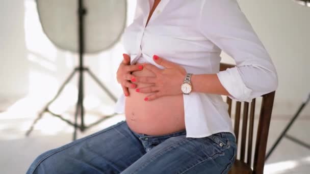 Feliz esperando mulher em um estúdio branco com luzes equipamentos vestindo camisa branca e jeans massageando sua barriga. Jovem mulher se preparando para se tornar uma mulher mãe. Fecha. Prores 422 — Vídeo de Stock