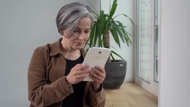 Mujer leyendo noticias usando tableta digital. Mujer madura en chaqueta marrón y jeans sostiene tableta de pie cerca de la ventana. Prores 422 — Vídeos de Stock
