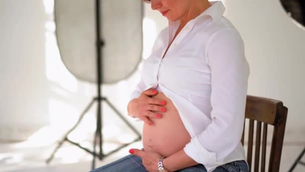 Close up on belly. Beautiful pregnant woman sitting in the studio with lights equipment wearing white shirt massaging her belly. Young getting ready to become a mother woman. Prores 422 — Stock Video