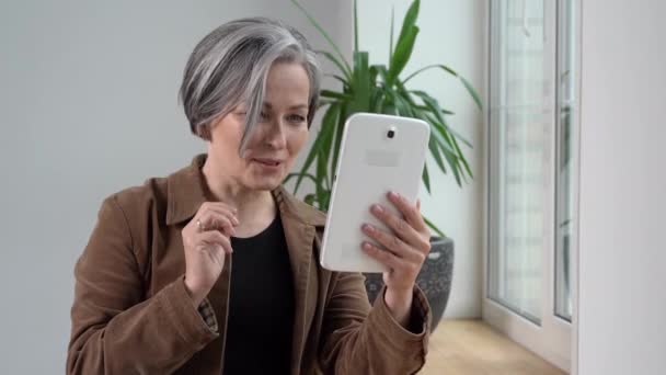 Mujer hablando con un nieto utilice tableta digital para videollamada sonriendo a la cámara. Mujer madura en chaqueta marrón y jeans sostiene tableta de pie cerca de la ventana. Prores 422 — Vídeos de Stock