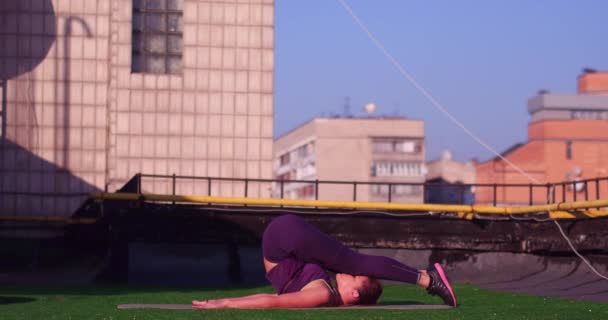 Fille brune sportive portant des vêtements de sport violets, faisant des exercices d'étirement sur le toit. Slim fille sportive effectue des exercices d'étirement sur le toit du bâtiment, fond urbain. Prores 422 — Video
