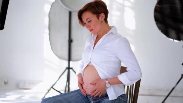Bela mulher grávida sentada no estúdio com luzes equipamentos vestindo camisa branca massageando sua barriga. Jovem se preparando para se tornar uma mulher mãe. Prores 422 — Vídeo de Stock