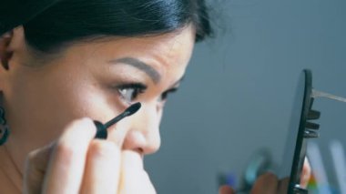 Kadın alt kirpiklerini rimel ile boyuyor. Bir elinde ayna, diğer elinde makyaj malzemesi tutuyor. Makyaj konseptini uyguluyorum. Yüksek kalite 4k görüntü