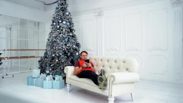 Un hombre de mediana edad se sienta en un sofá con un teléfono cerca de un árbol de Navidad. Cuarentena de Año Nuevo. Interior de Navidad. Vacaciones en casa — Vídeos de Stock